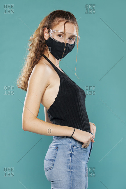 Side view of female wearing medical mask and plastic safety goggles standing on blue background in studio and looking at camera