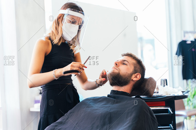 Barber woman in protective mask and face shield shaving male with sharp straight razor while working in barbershop during coronavirus outbreak