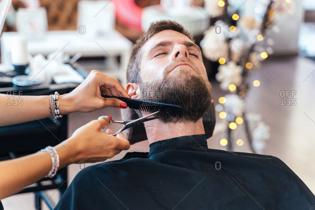 Cropped unrecognizable professional female barber cutting beard on male customer with scissors while working in modern barbershop