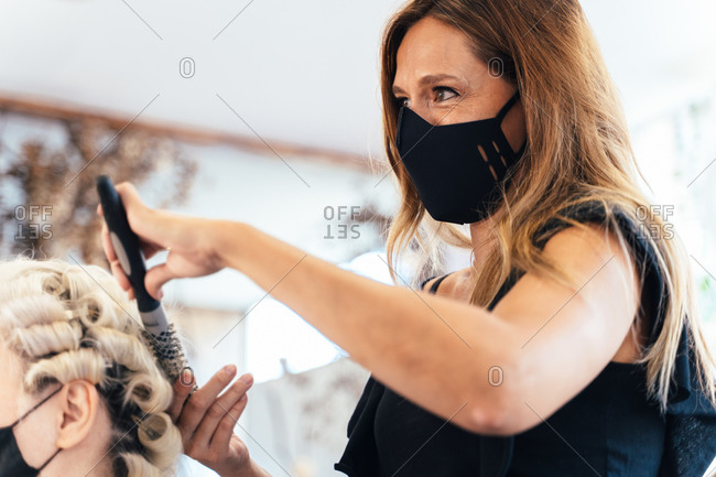Side view of female stylist in protective mask drying hair of cropped unrecognizable woman sitting in front of mirror in beauty salon