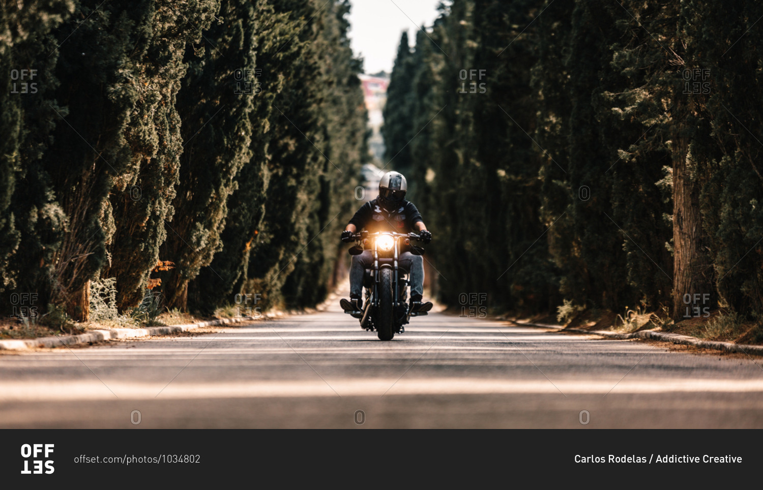 Biker in black leather jackets and helmet riding powerful motorcycle on asphalt road leading between green forest in countryside
