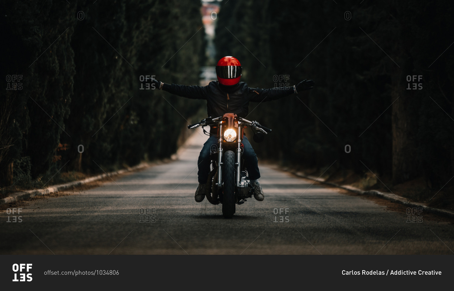Biker in black leather jackets and helmet with open arms riding powerful motorcycle on asphalt road leading between green forest in countryside