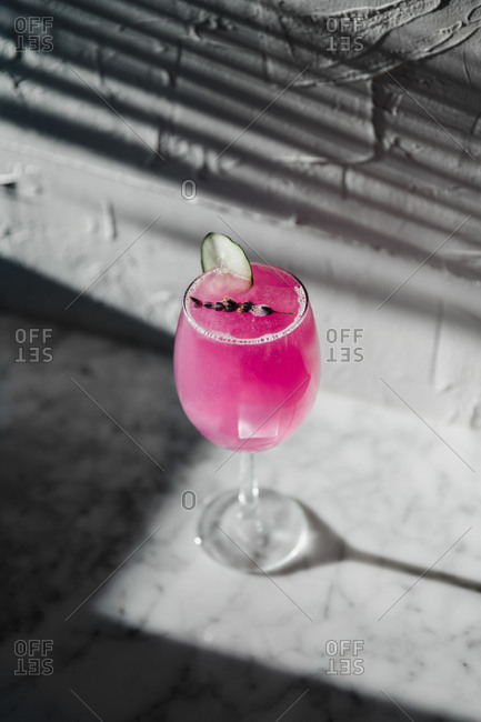 From above of wineglass of refreshing bright purple cocktail garnished with herbs and sliced lime placed in sunlight against blurred gray background