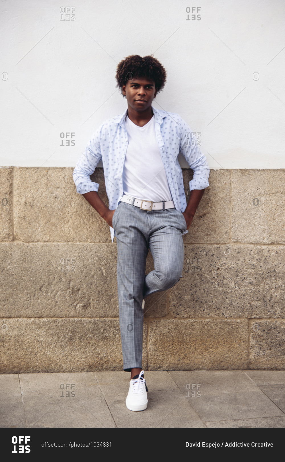 Determined African American male in trendy outfit and with Afro hairstyle leaning on stone wall of building and looking away