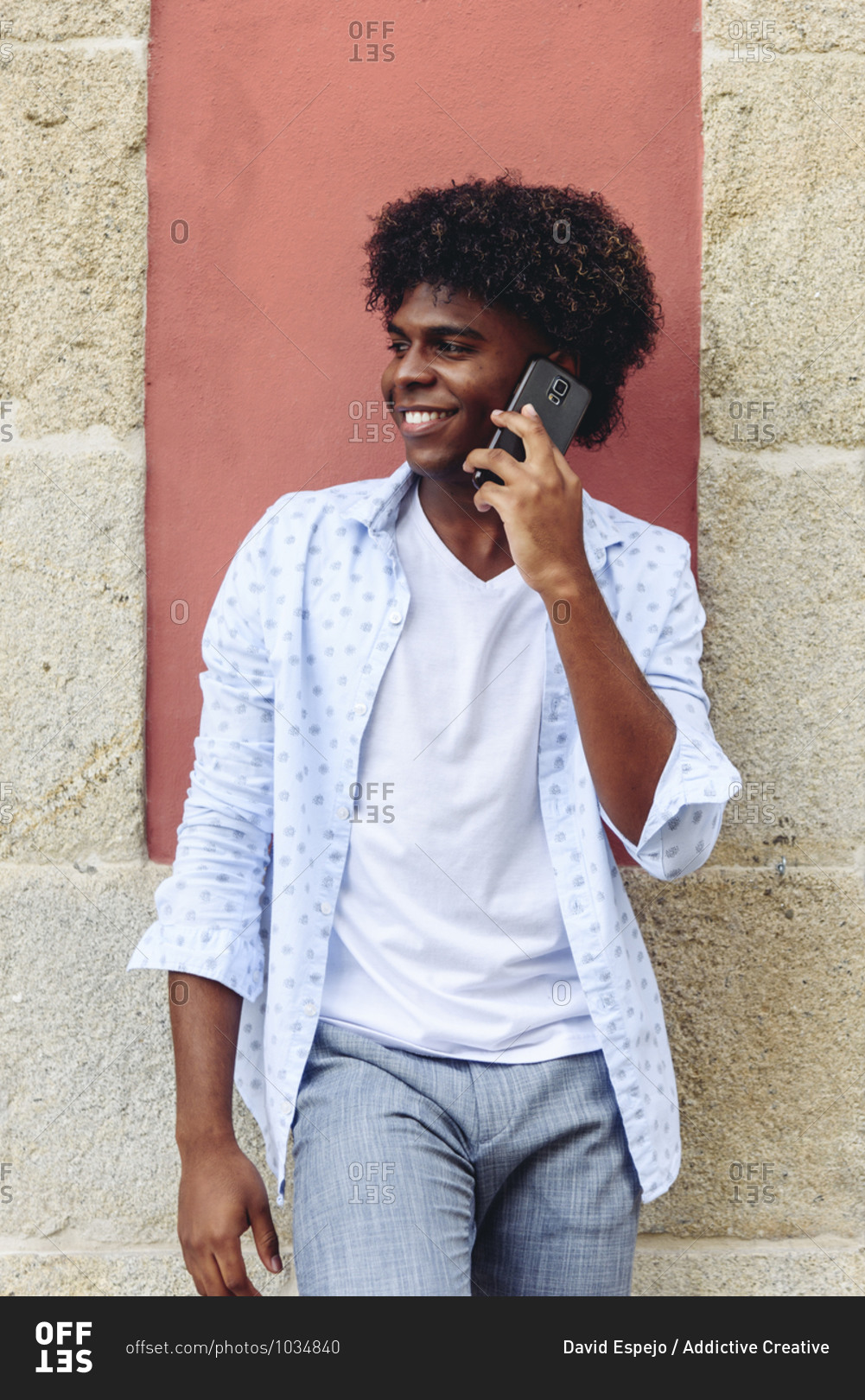 Positive African American male in stylish summer outfit leaning on building in city and speaking on smartphone while looking away