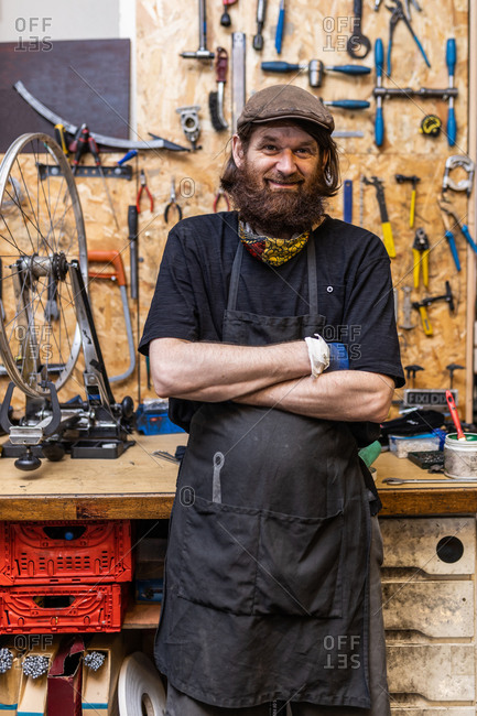 Smiling middle aged bearded craftsman in workwear looking at camera friendly while standing against weathered workbench with various tools in bicycle service workshop