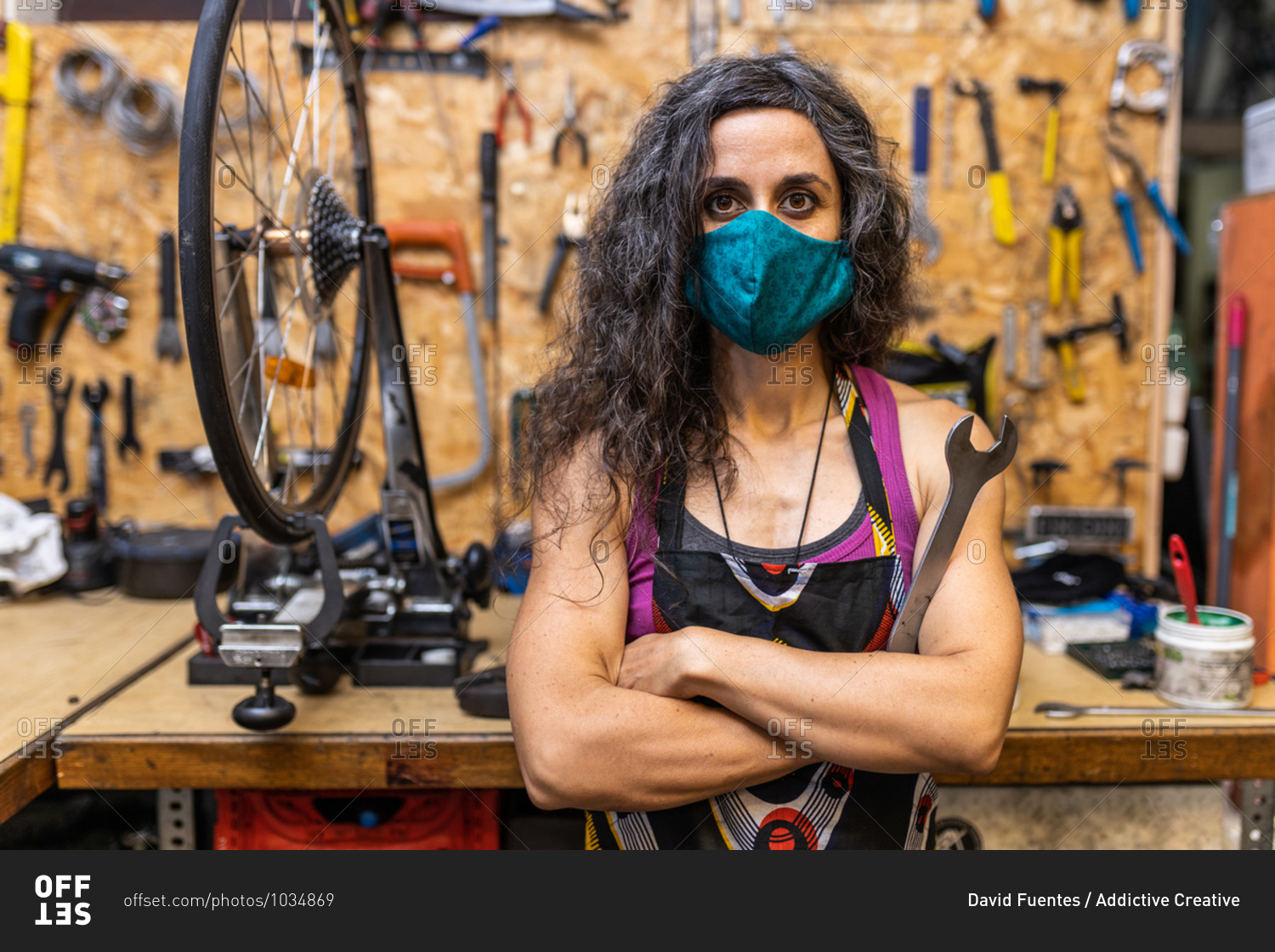 Serious adult ethnic female mechanic in workwear and protective mask holding wrench and looking at camera while standing against weathered workbench with various tools in bicycle service workshop