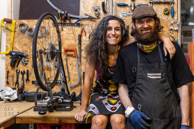Positive ethnic couple of owners of bicycle service workshop looking at camera and smiling friendly against weathered wall with various repair instruments
