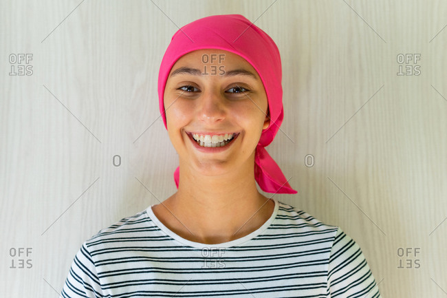 Young delighted female with cancer wearing headscarf standing in apartment and looking at camera