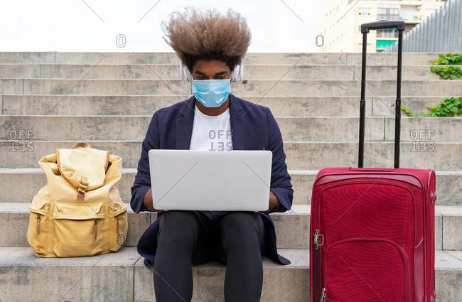 Black male entrepreneur with afro and wearing a protective face mask in suit sitting on stairs in city and working on project while using laptop and listen to music on headphone