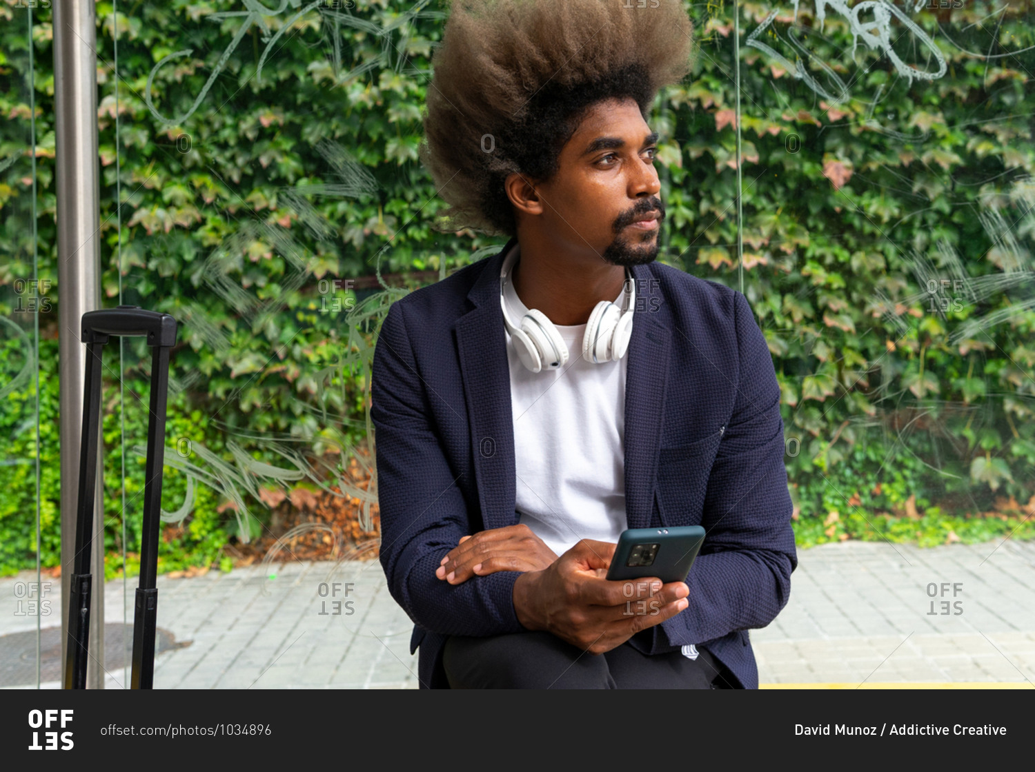 African man with afro hair sitting on a bench outside with headphones around his neck and a suitcase while using his mobile