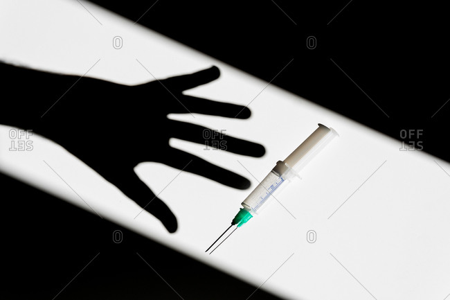 Shadow of crop anonymous person reaching out for syringe placed on white table