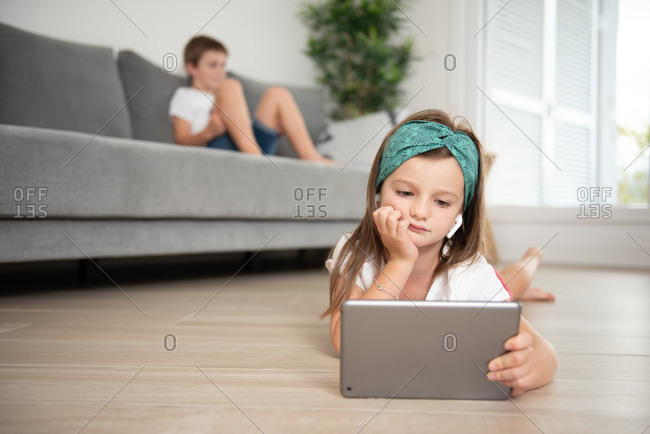 Little girl with true wireless earphones lying down on the floor while entertained using tablet during weekend