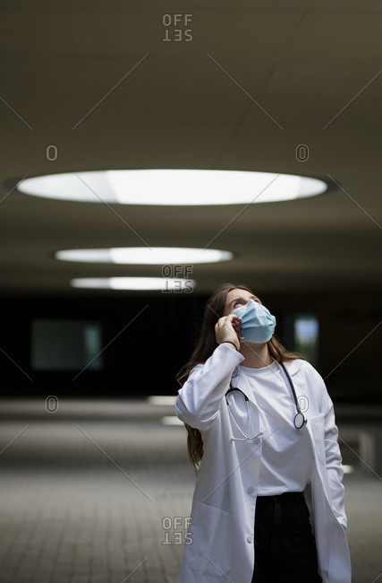 Young female medical practitioner in white coat and protective mask with stethoscope talking on mobile phone in hospital hall