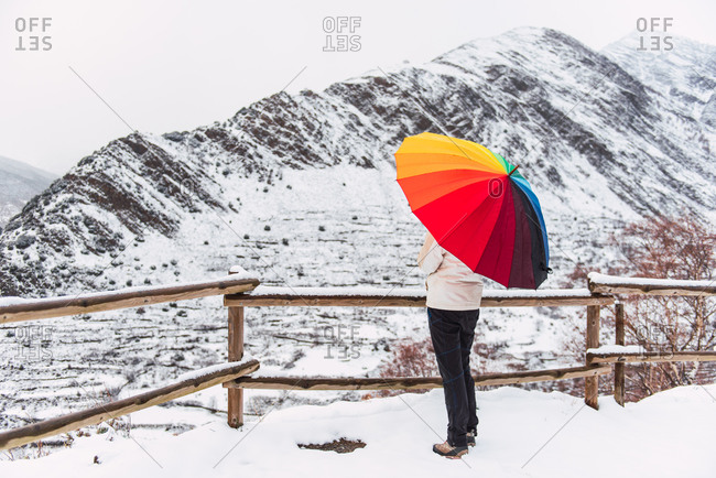Side view of anonymous person standing under colorful umbrella and enjoying amazing view of Pyrenees mountain ridge covered with snow in winter