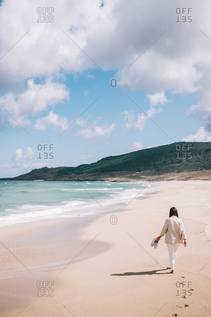 Full body back view of unrecognizable female traveler walking barefoot on empty sandy beach near waving sea and enjoying fresh breeze during summer holidays in Spain