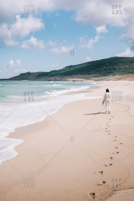 Full body back view of unrecognizable female traveler walking barefoot on empty sandy beach near waving sea and enjoying fresh breeze during summer holidays in Spain
