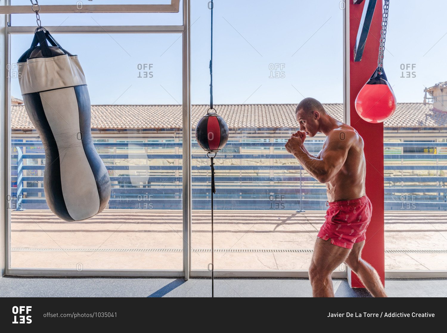 Side view of determined male boxer in boxing gloves punching heavy bag during intense workout in gym