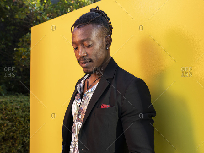Serious African American male model in fancy outfit standing near yellow wall on street and looking down