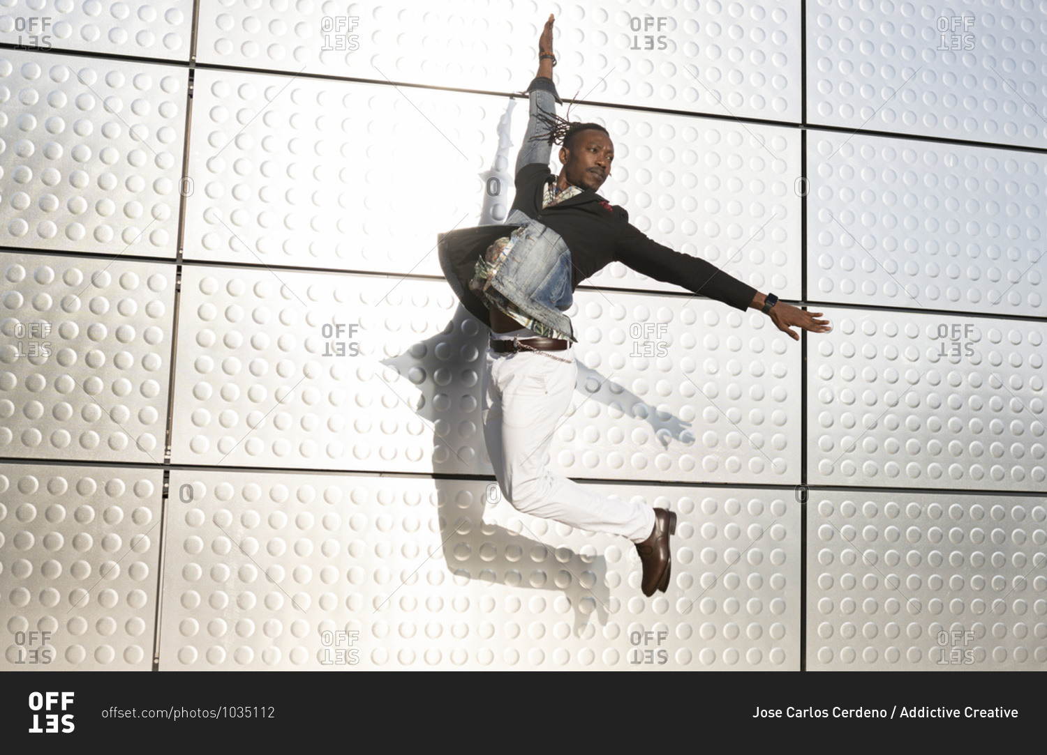 Side view of well dressed ethnic male entrepreneur jumping near urban building with outstretched arms and looking away