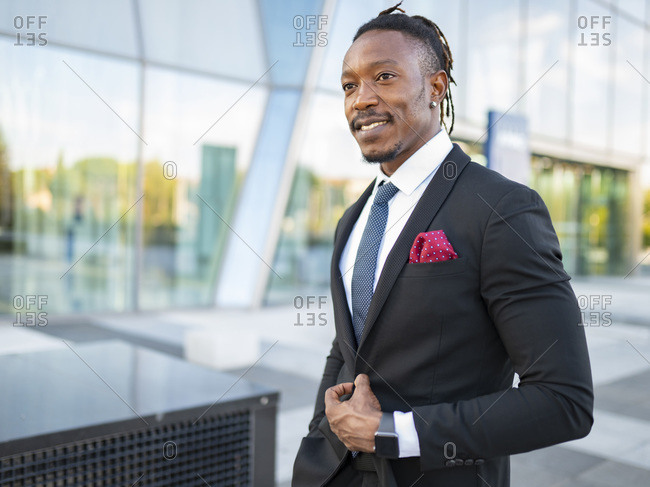 Cheerful African American male entrepreneur wearing elegant suit walking along glass office building in city center and looking away