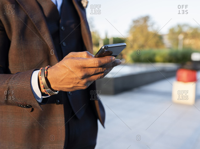 Unrecognizable ethnic businessman in stylish suit standing in city and surfing Internet on smartphone while working remotely on project