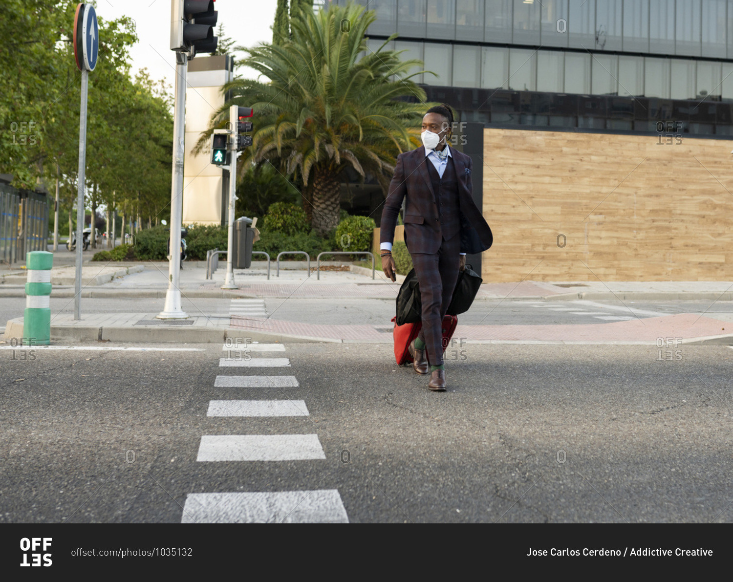 Serious black male entrepreneur wearing medical mask crossing road in city while walking with bags towards airport during coronavirus epidemic