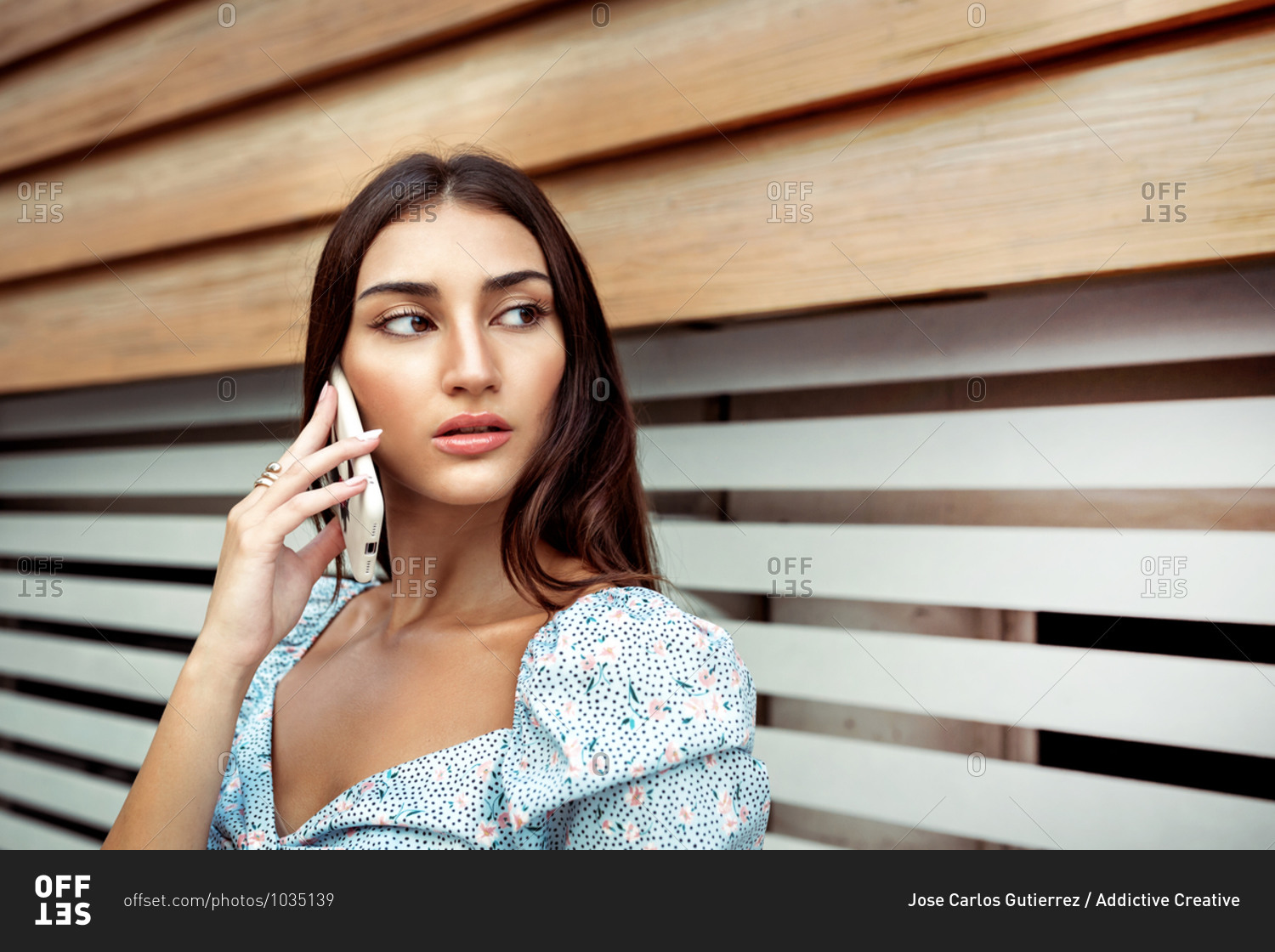 Young ethnic female in stylish blouse talking smartphone while standing near shuttered window of modern building