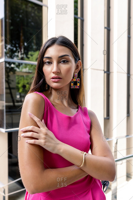 Young slim ethnic female in stylish pink overall with elegant earrings and bracelet standing against blue wall of contemporary building