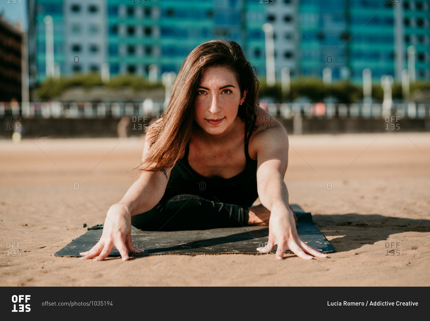 Smiling flexible female sitting on mat in Janu Sirsasana and doing forward bend while looking at camera and practicing yoga on beach in summer