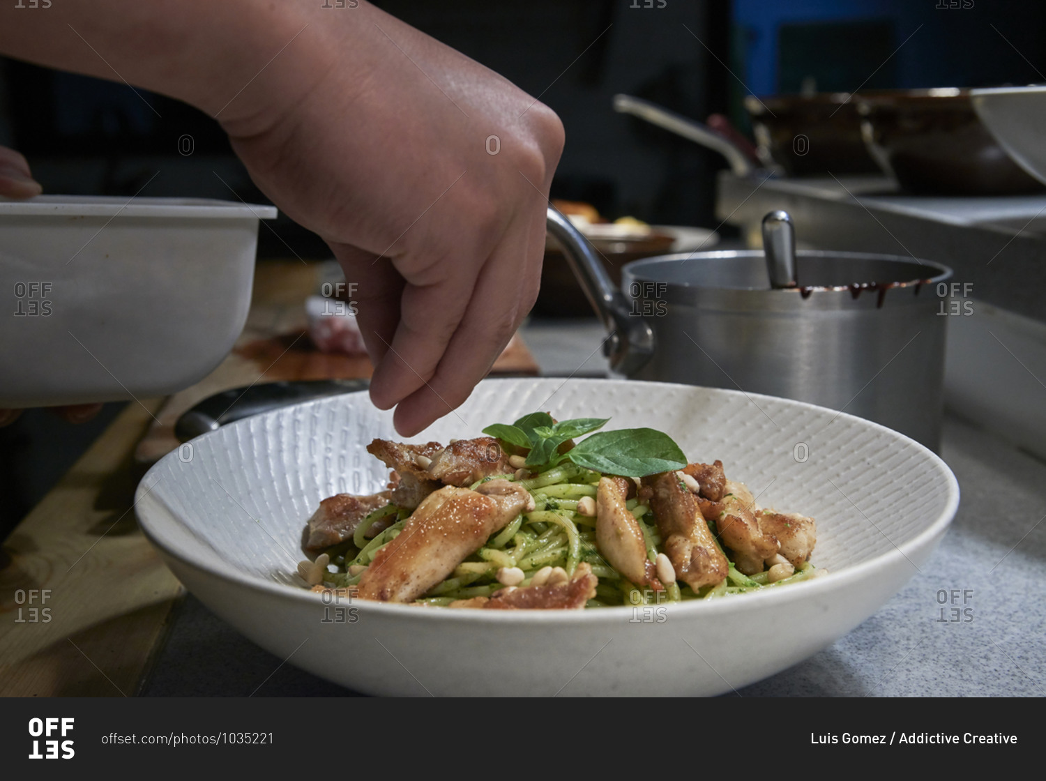 Closeup of crop anonymous cook seasoning and garnishing spaghetti with pesto sauce and chicken served in white bowl while working in restaurant kitchen