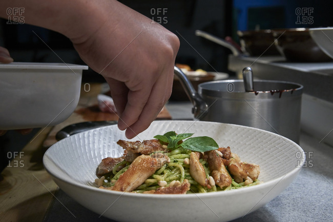Closeup of crop anonymous cook seasoning and garnishing spaghetti with pesto sauce and chicken served in white bowl while working in restaurant kitchen