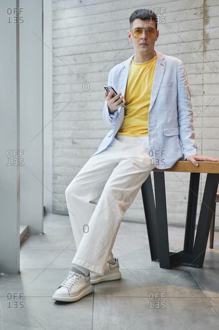 Serious male entrepreneur wearing stylish clothes leaning on table in bright workplace and browsing cellphone
