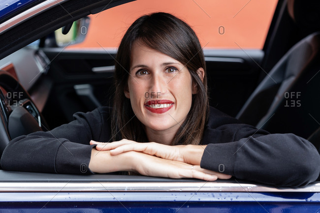 Cheerful female driver leaning on automobile while sitting in car and looking at camera