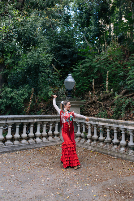 Full body graceful young female in typical elegant red gown performing Flamenco dance with arm raised on terrace with aged fence against green trees