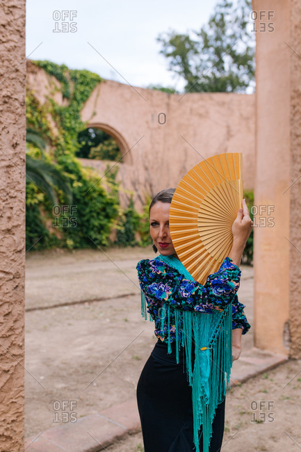 Gorgeous female Flamenco dancer in colorful garment covering half of face with open fan while standing against blurred old stone wall with arched passage