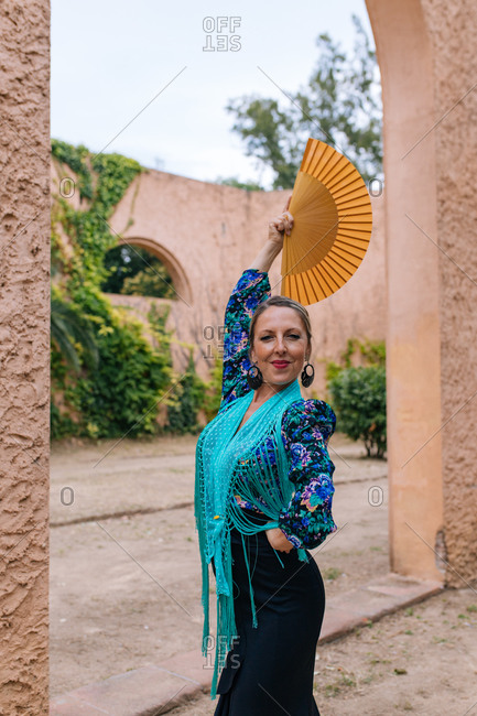 Gorgeous female Flamenco dancer in colorful garment with open fan while standing against blurred old stone wall with arched passage