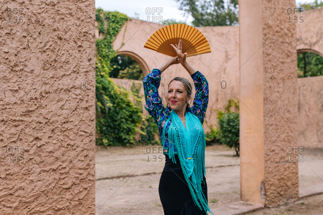 Gorgeous female Flamenco dancer in colorful garment with open fan while standing against blurred old stone wall with arched passage