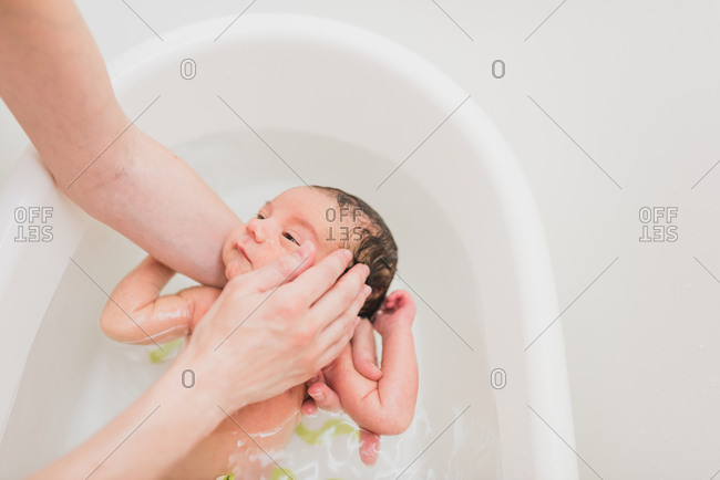 From above unrecognizable parent washing and touching face of crying newborn baby in warm water in basin at home