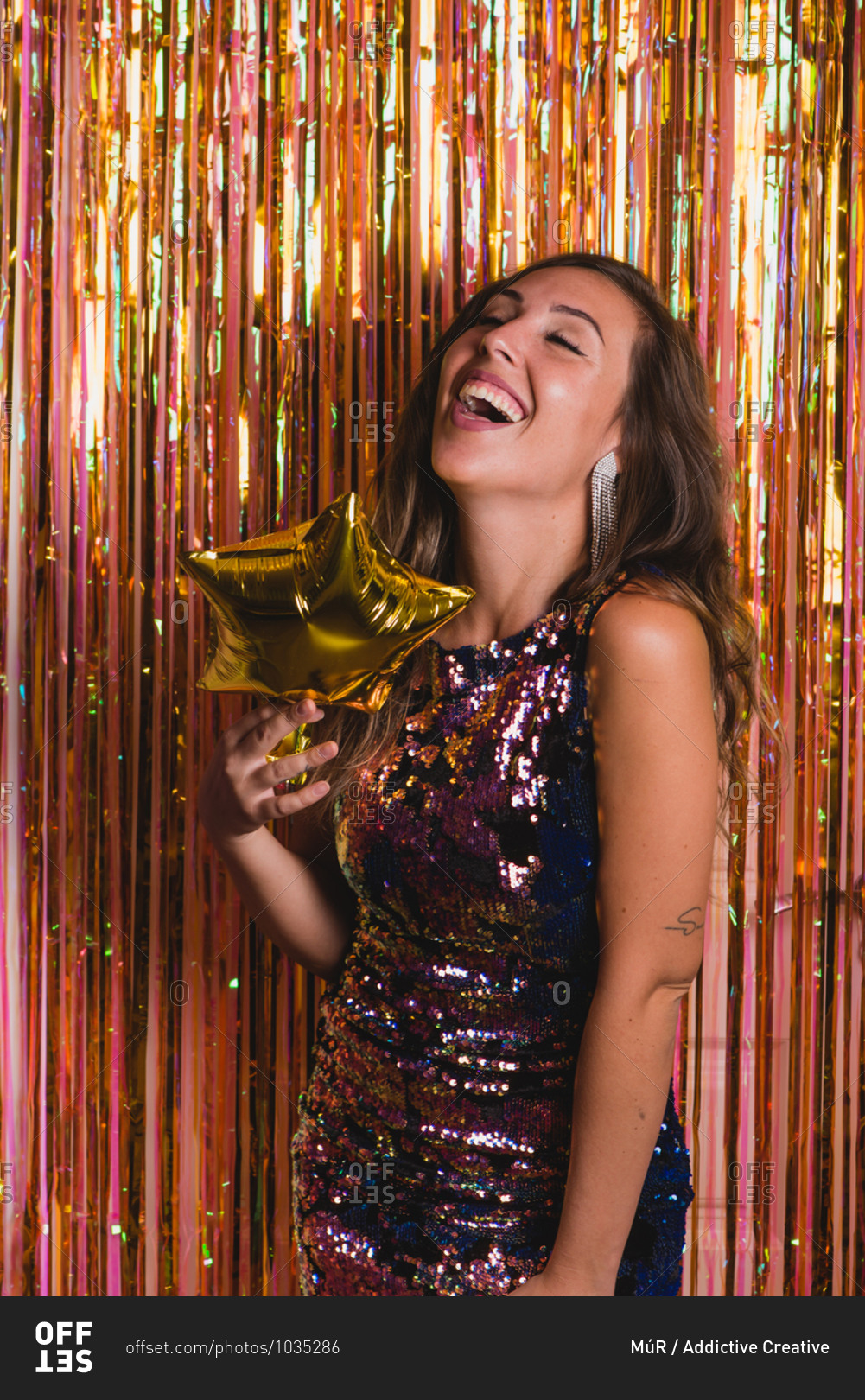 Excited female in sparkling glamour dress standing with golden star on background of foil tinsel and laughing with closed eyes during party celebration