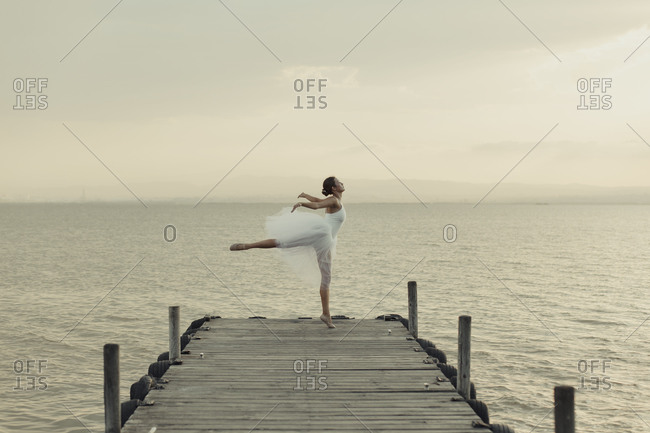 Full body young graceful female dancer in white gown standing on tiptoes with leg raised while performing ballet movements on wooden boardwalk against sea in summer evening