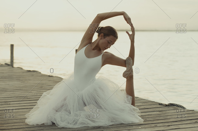 Full body flexible classic ballet female dancer in elegant white dress performing sensual pose on wooden pier against blurred sea in summer evening