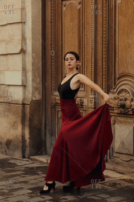 Charming young Hispanic brunette in red skirt performing traditional Flamenco dance near wooden door of old building