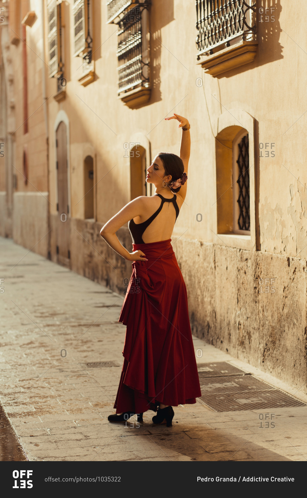 Full body back view of young Hispanic female dancer in traditional outfit performing Flamenco dance on old street with aged stone buildings