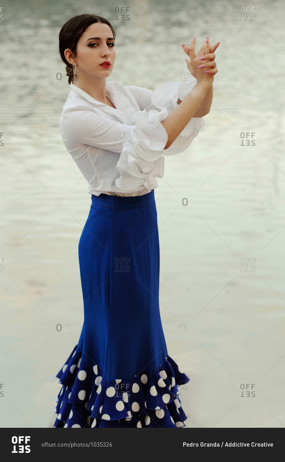 Side view of sensual young Hispanic woman in elegant white blouse and blue skirt performing graceful movements with hands during Flamenco dance against lake water