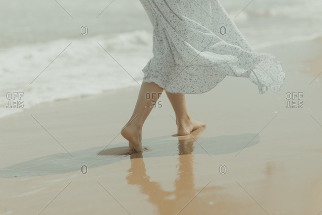 Back view of crop unrecognizable barefoot female in white summer dress walking alone on sandy beach in sunny summer day in Spain