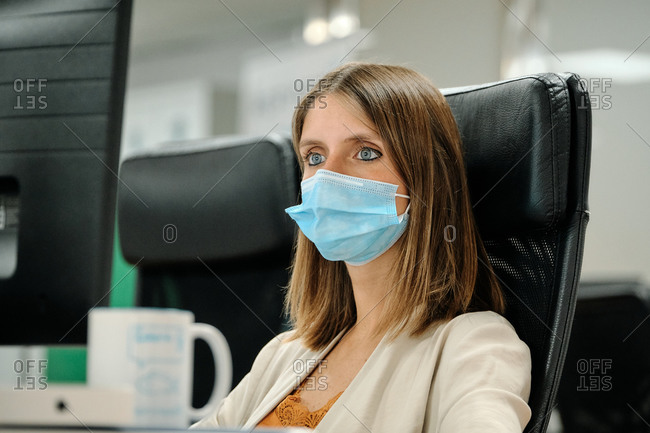 Focused female office employee in protective mask using computer while working in modern office during coronavirus pandemic
