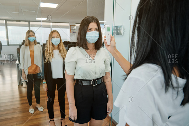 Receptionist with thermometer gun checking body temperature of coworkers before entering office space during coronavirus pandemic