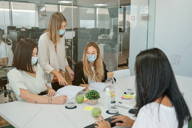 Young female manager in medical mask explaining business idea to coworkers while standing together in modern workspace and discussing project