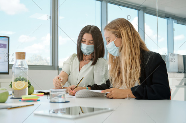 Positive young female colleagues in medical masks for coronavirus prevention taking notes in notebook and discussing details of business project while having meeting in contemporary workspace